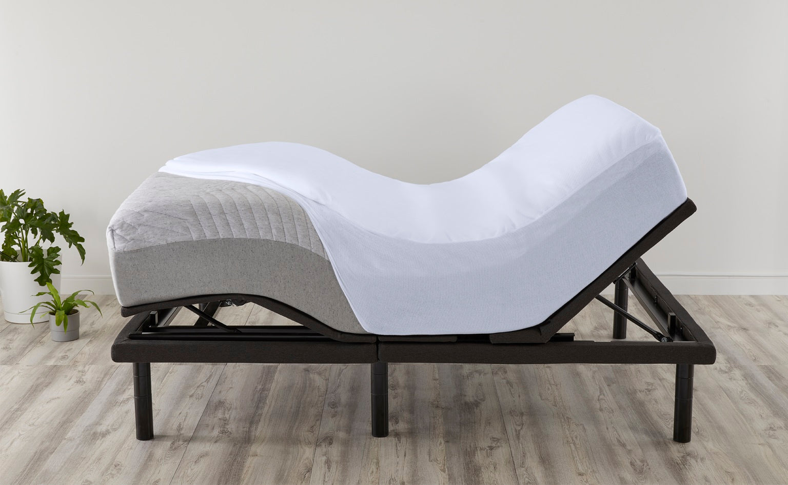 A white mattress on an adjustable bed frame is shown in an undulating, zero-gravity position. The setup is in a bright room with light wood floors and two small potted plants in the background.