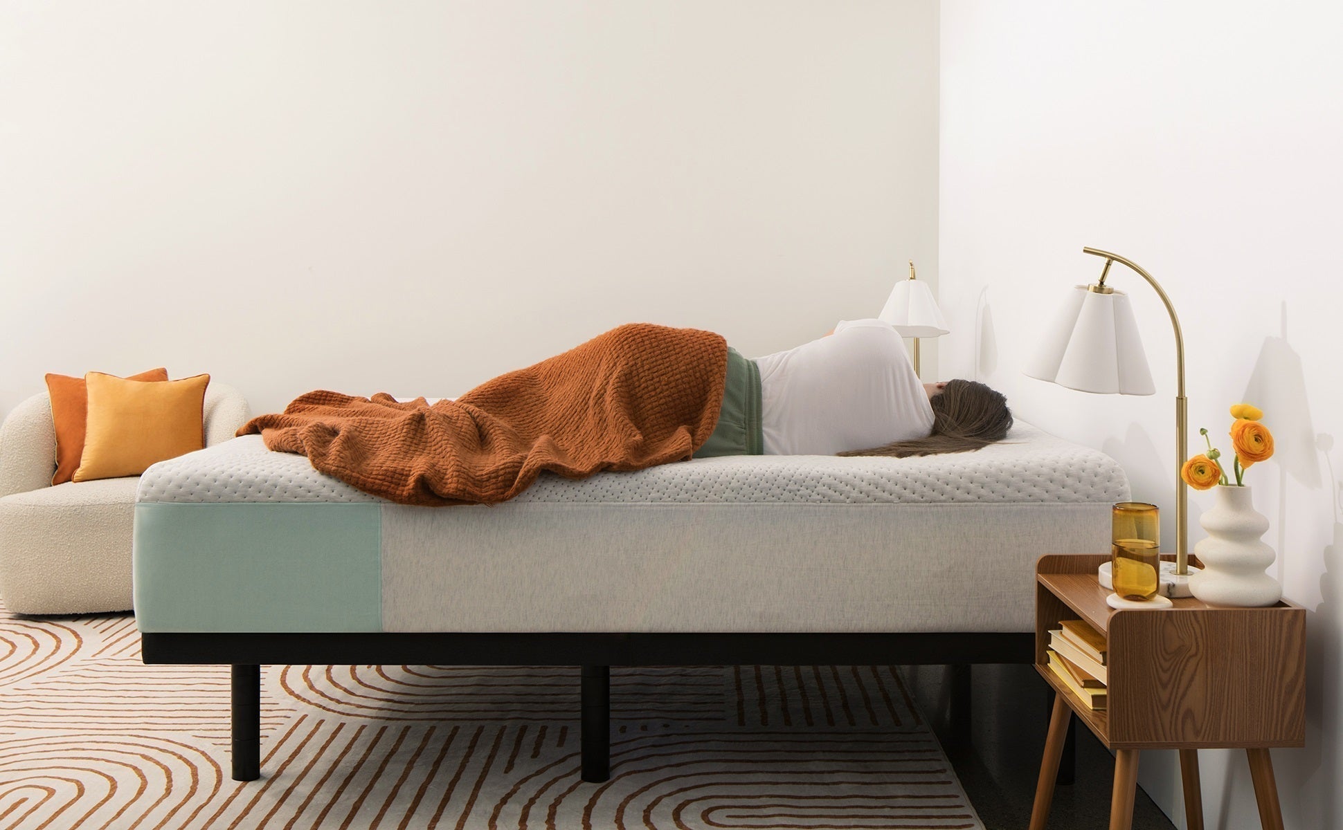 Young woman sleeping on their side on a memory foam mattress with a brown blanket in a cozy, modern bedroom featuring warm tones and minimalist decor.