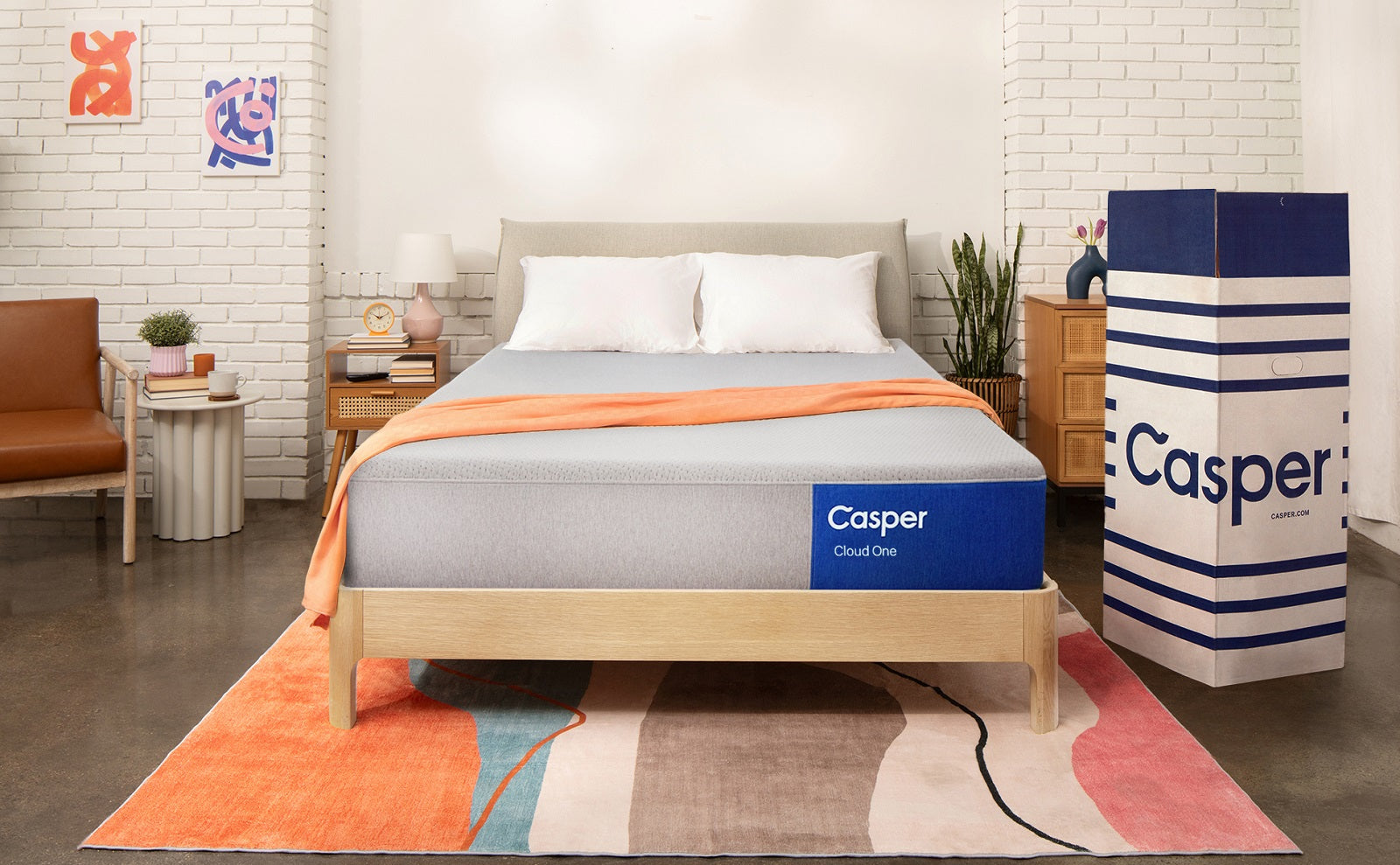 A Casper Cloud One mattress rests on a light wood bed frame in a modern bedroom setting, positioned next to its tall, branded shipping box. The room features white brick walls, a colorful orange-toned rug, and a cozy seating area.