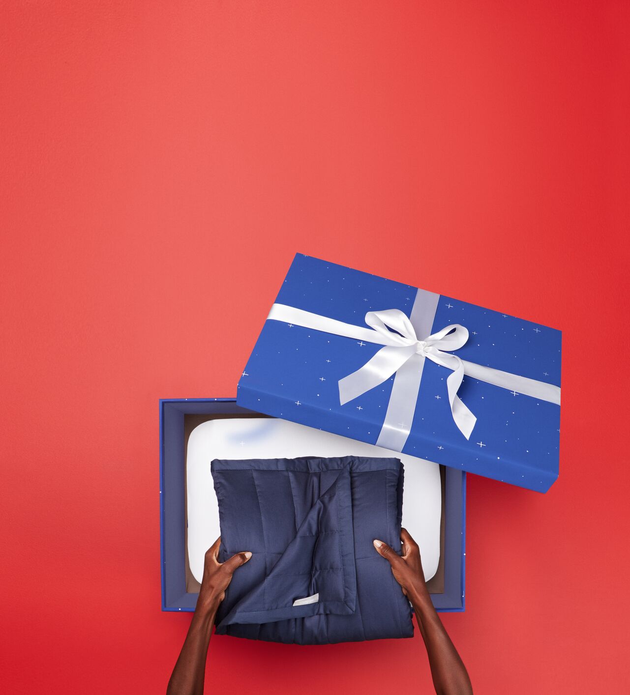 Hands place a folded navy blanket into an open blue gift box with a white ribbon, set against a red background.