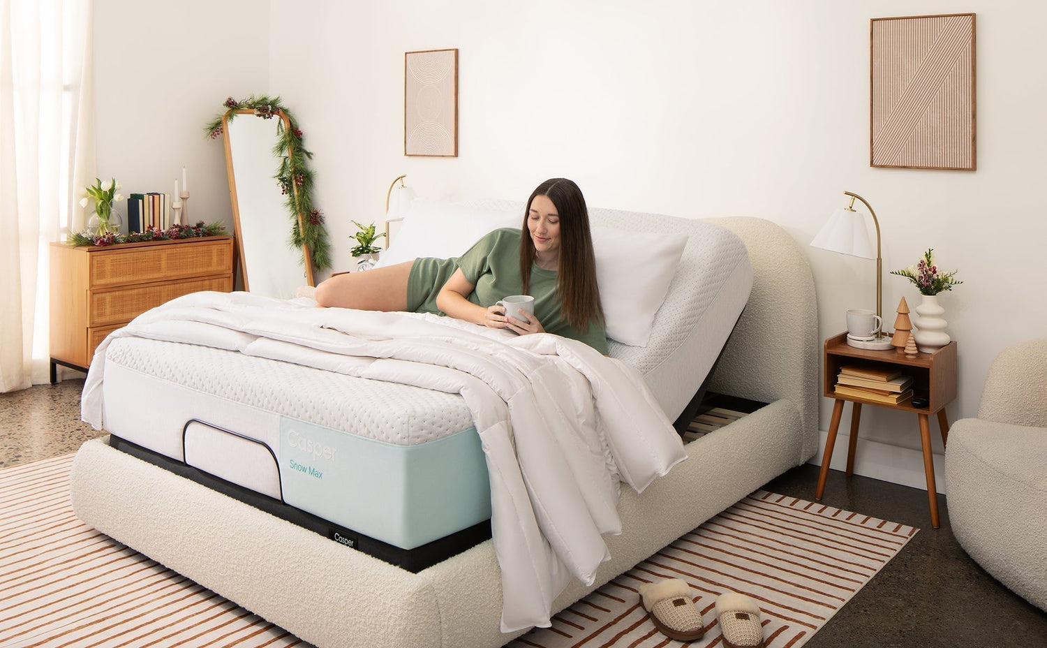 A woman relaxes on a Casper Snow Max mattress in an adjustable bed, holding a mug in a cozy, modern bedroom with neutral tones and holiday decor.