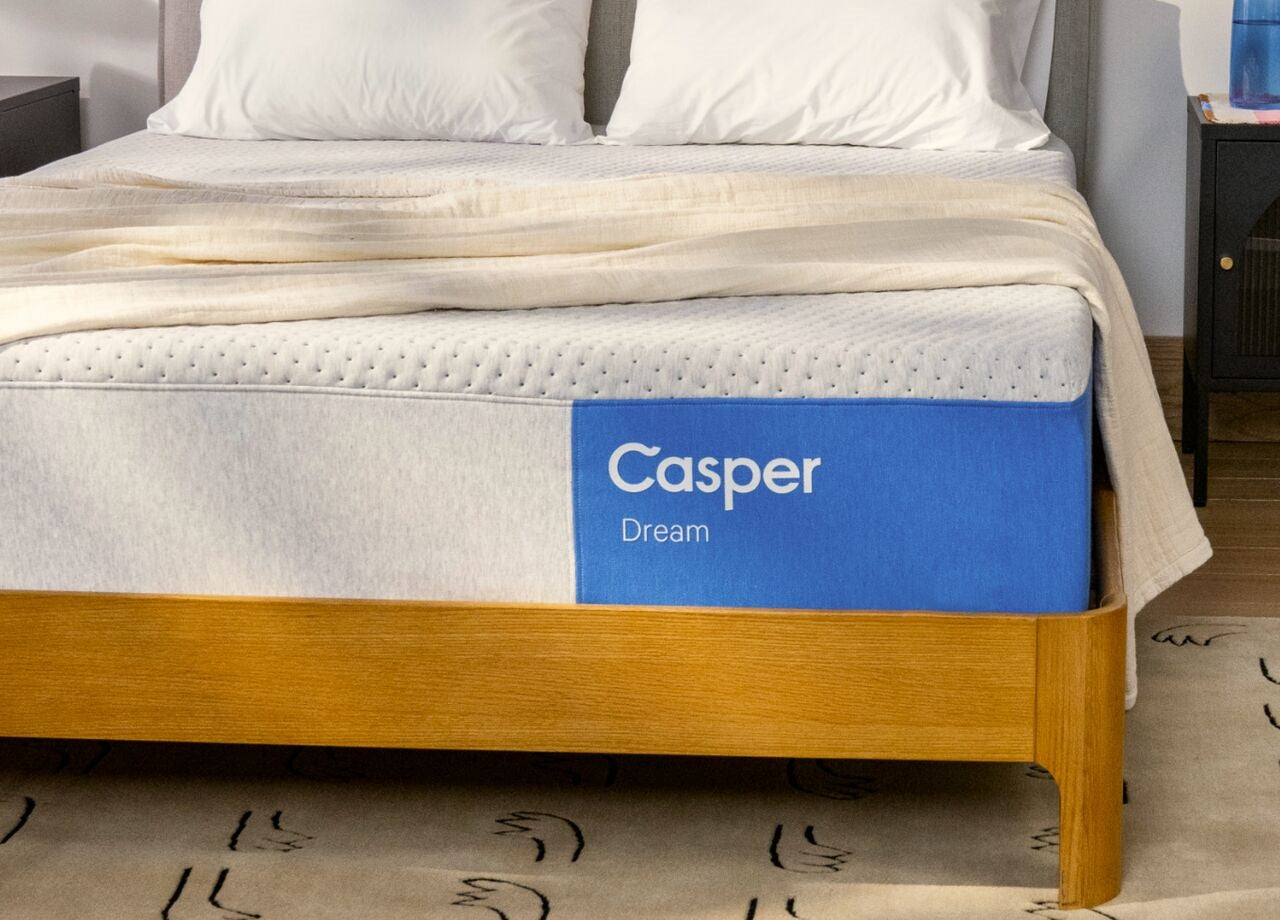 Casper Dream mattress on a wooden bed frame with white bedding and a beige throw, showing the blue side panel branding.