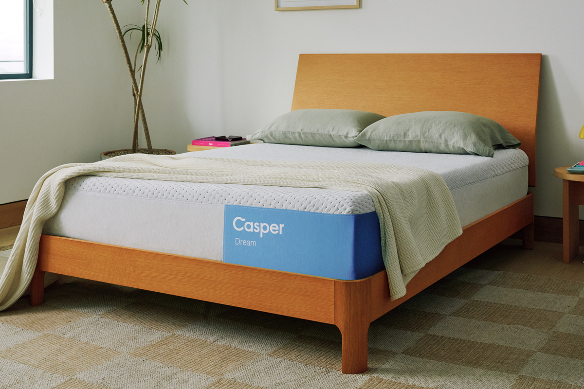 A modern wooden queen-size bed frame with a Casper Dream mattress, styled with green pillows and a neutral throw blanket in a brightly lit room.