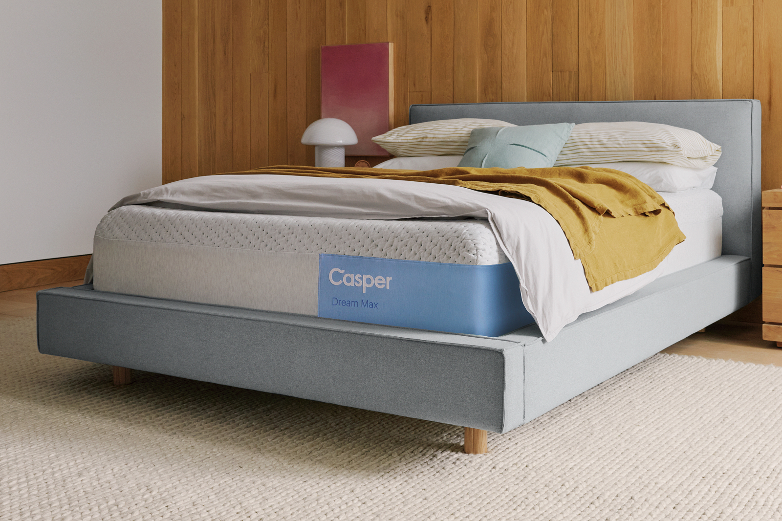 A Casper Dream Max mattress made with crisp white linens and a mustard-yellow throw blanket, resting on a light blue upholstered bed frame in a modern bedroom with warm wood paneling.