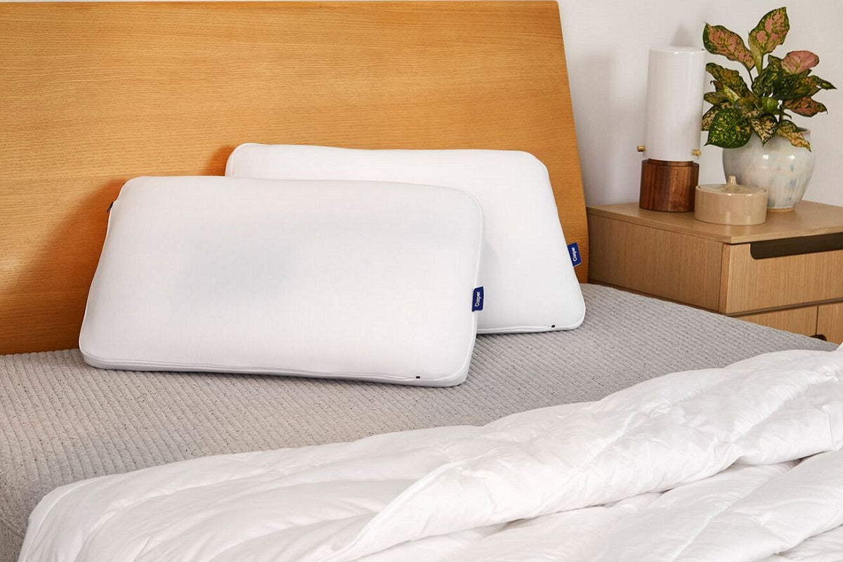 A photograph of two white Casper pillows propped against a wooden headboard on a bed with a gray textured cover and white duvet.