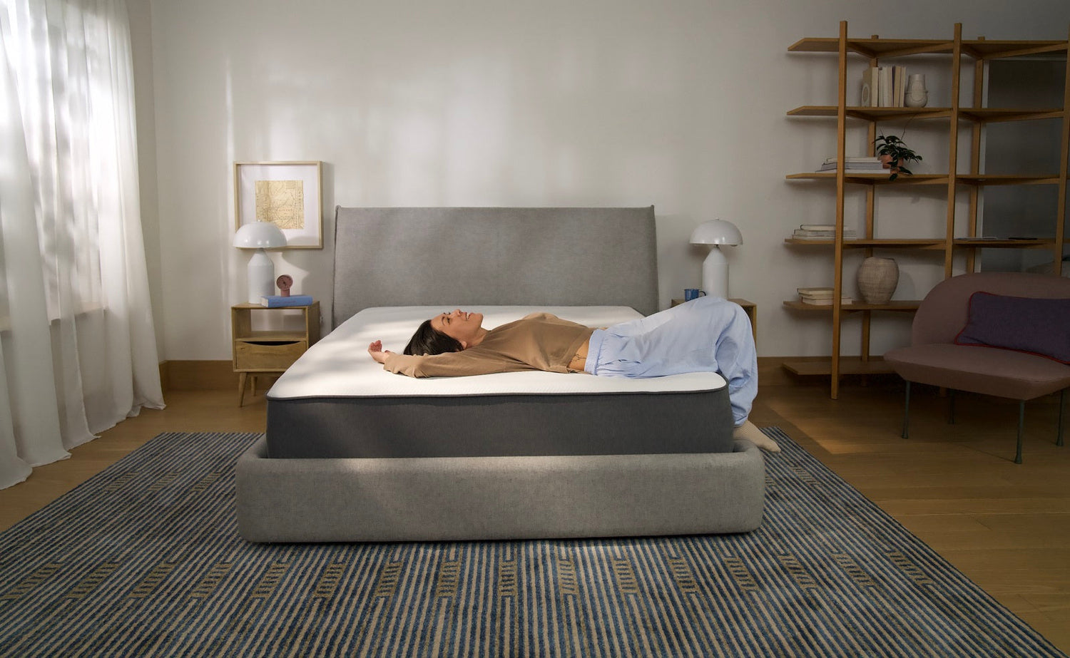 Person lying on their back across a modern bed, relaxing and testing the comfort of a mattress in a sunlit bedroom.