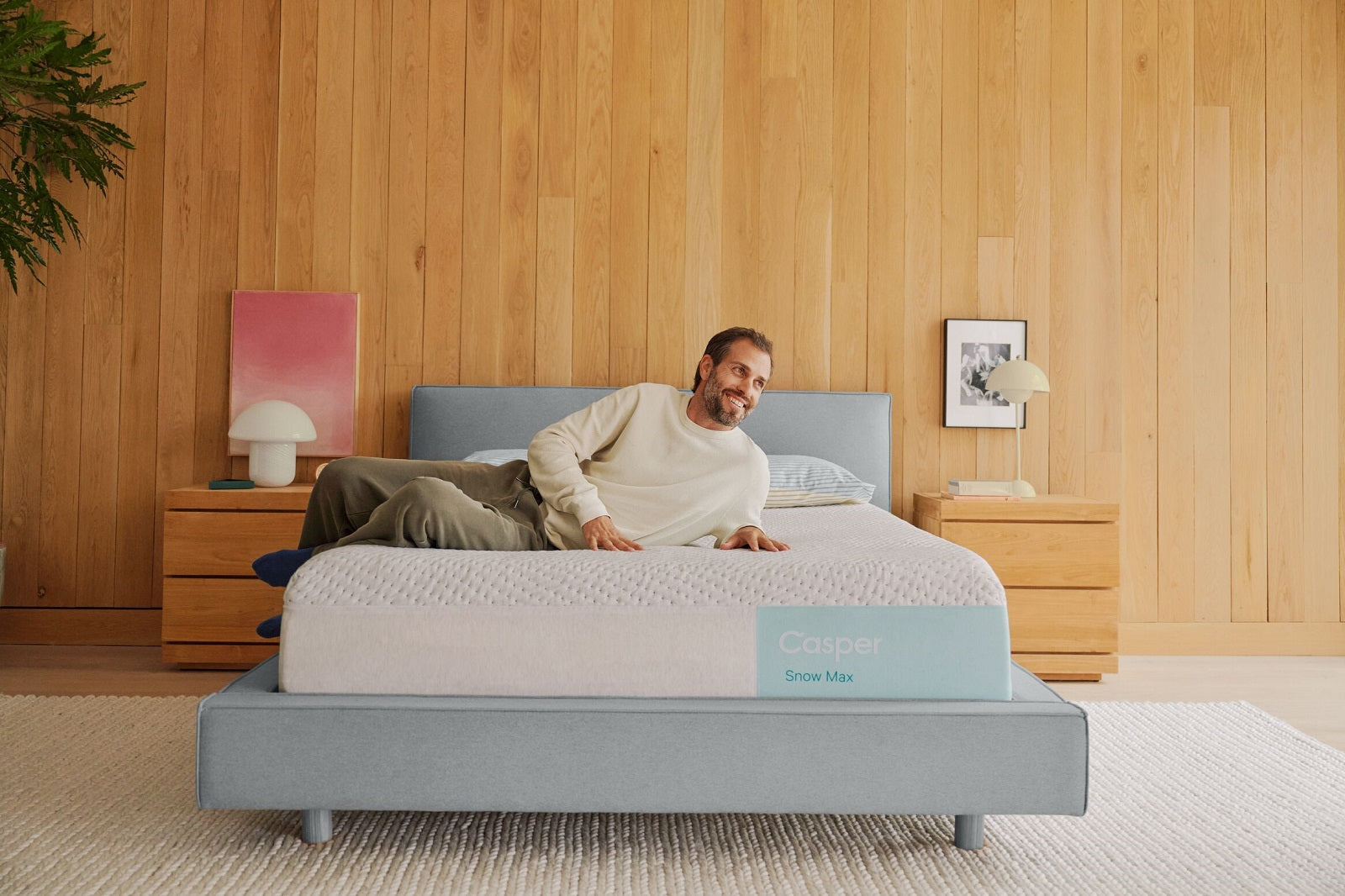 A man lies smiling on a Casper Snow Max mattress set on a light blue bed frame in a modern, wood-paneled bedroom.