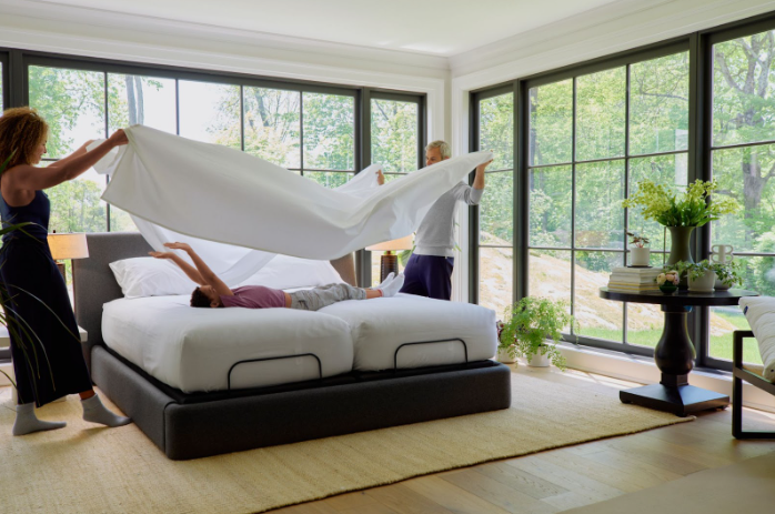 Two adults billow a white sheet over a split king adjustable bed in a bright room with floor-to-ceiling windows