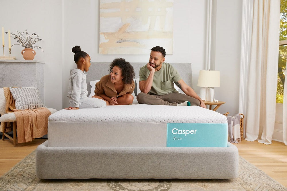 A family, consisting of two adults and one child, sitting and talking happily on a Casper Snow mattress in a bright, modern bedroom.