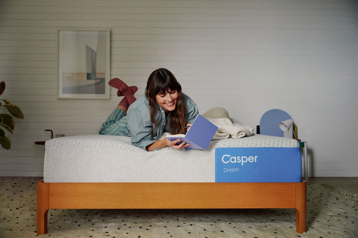 Woman in striped pajamas lies on a Casper Dream mattress, smiling while reading a blue book in a cozy bedroom.
