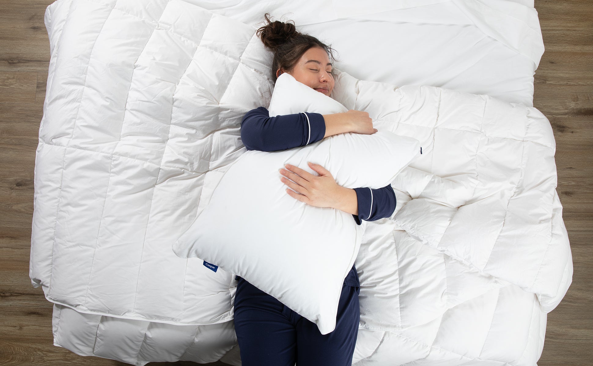 A woman in navy pajamas lies on a bed hugging a white Casper pillow, surrounded by soft white bedding.