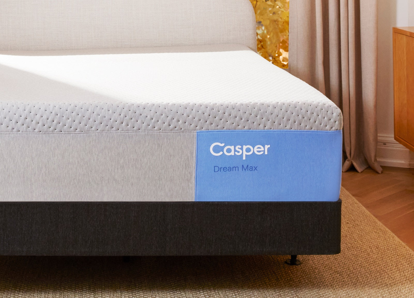 A close-up of a Casper Dream Max mattress corner on a dark gray upholstered frame, highlighting its textured white top and blue side panel branding.