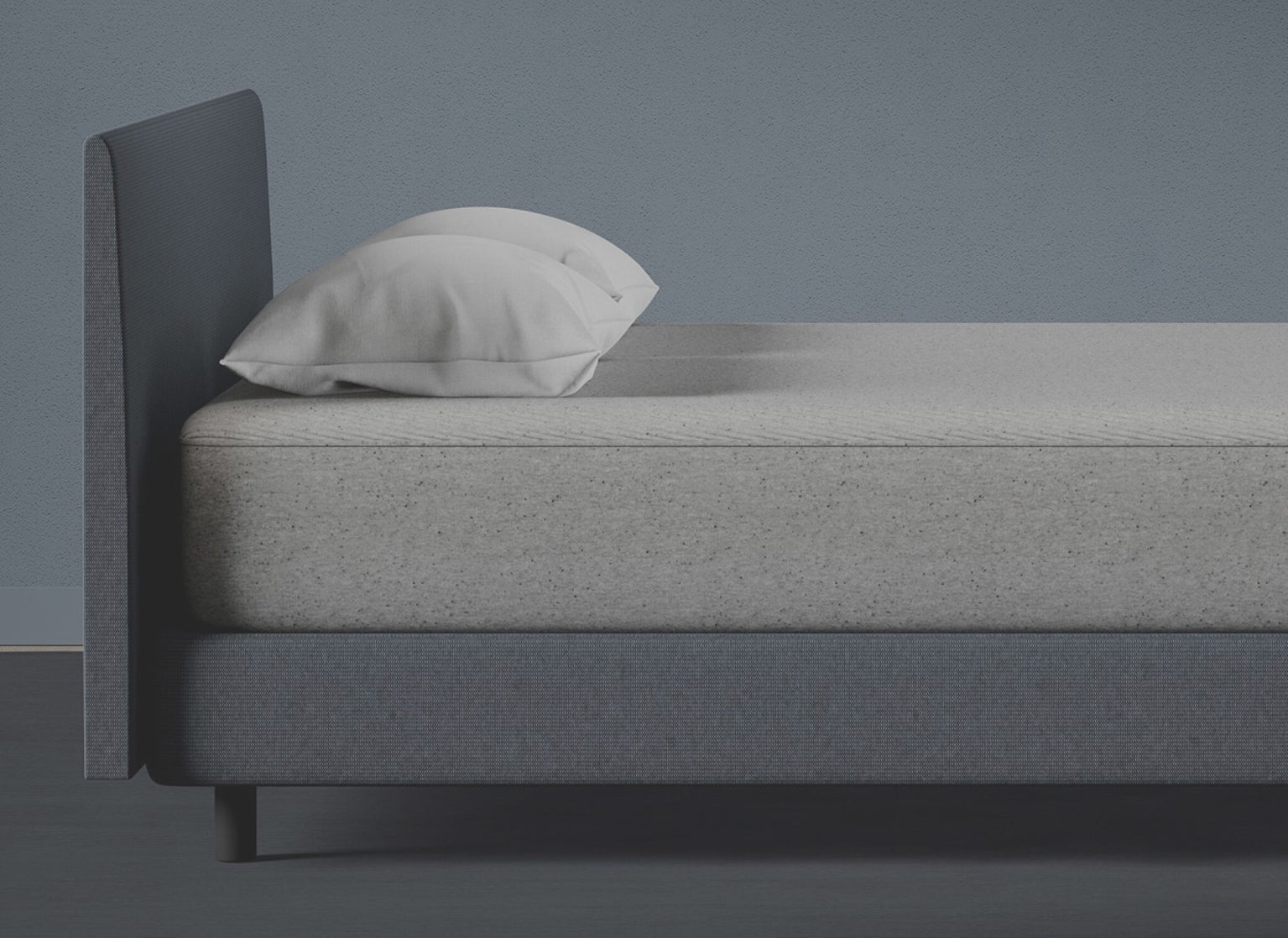A photograph of a Casper mattress with two white pillows on a dark gray upholstered bed frame against a plain gray wall.