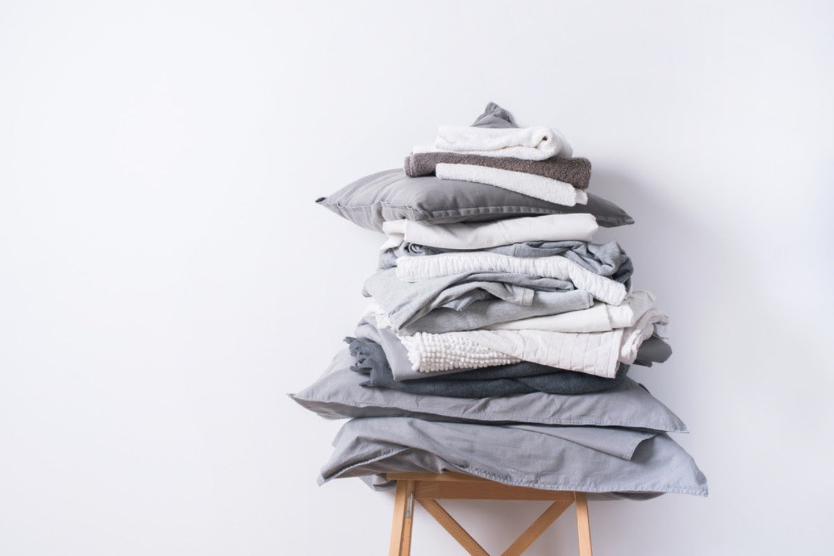 Tall stack of folded bed sheets, pillowcases, and towels on a wooden stool against a white background.