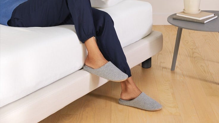 A person sitting towards the edge of their bed with their legs hanging off. One foot rests on the edge of the Upholstered Bed Frame.