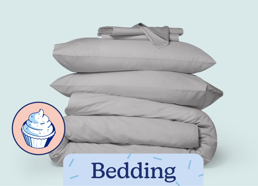 A stack of folded sheets sits on top of two pillows and a folded duvet against a white background. The text 'Bedding' is on the image with an illustration of a cupcake.