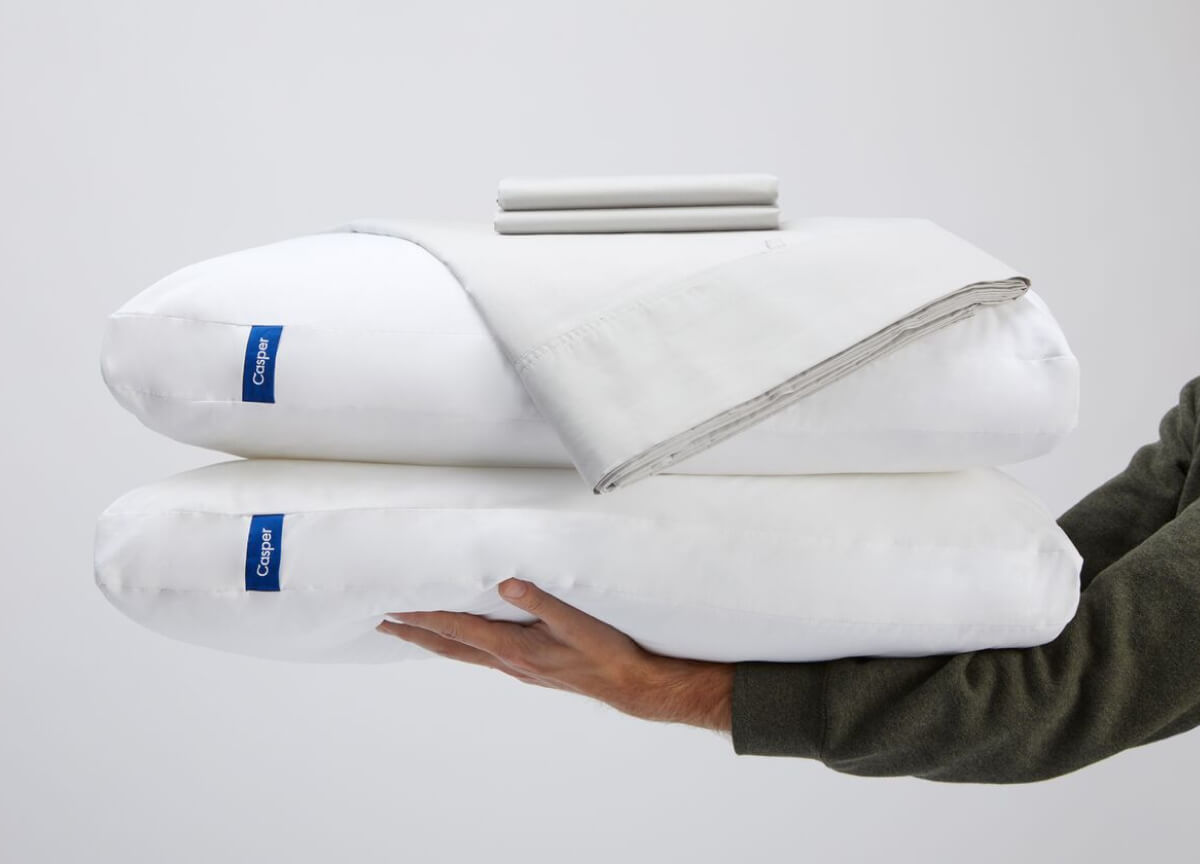 A person holds a stack of two pillows and a folded sheet set in their arms.