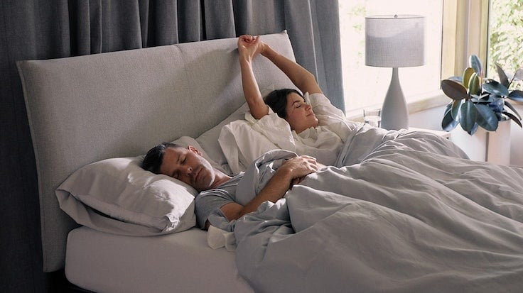 A couple lying in bed one morning. One of them is waking up and stretching.