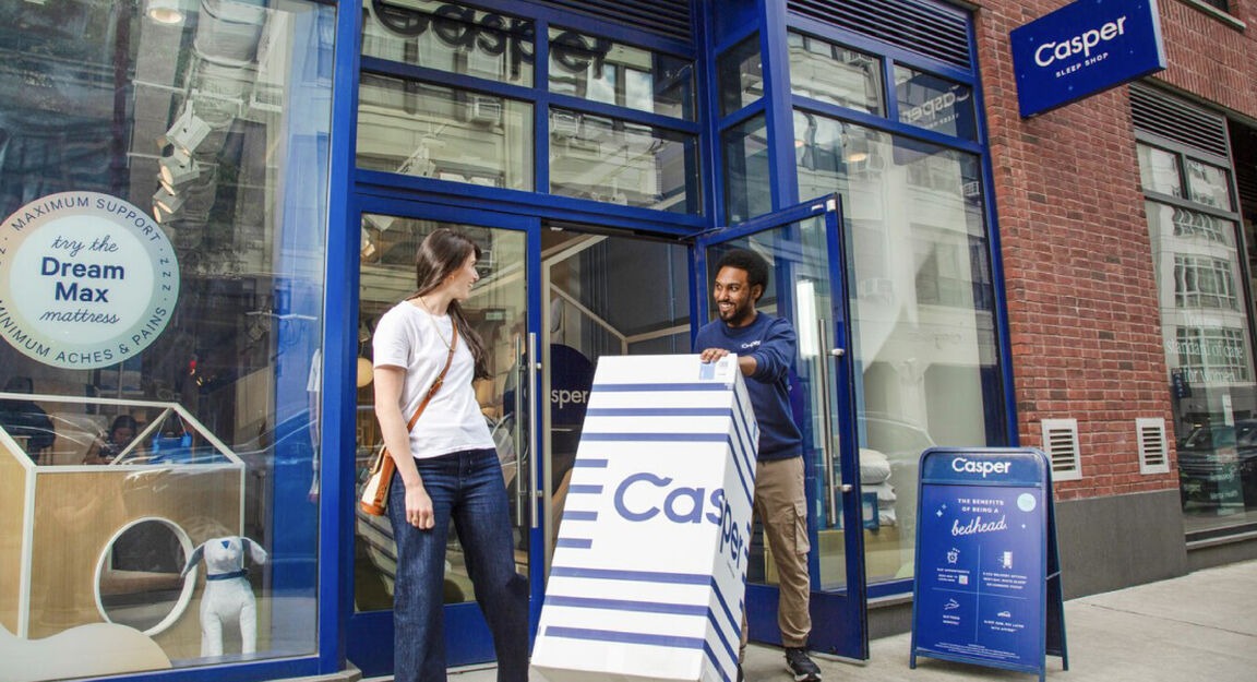 Photo outside a Casper retail store. The store associate is helping a customer wheel out her new Casper mattress in a box.