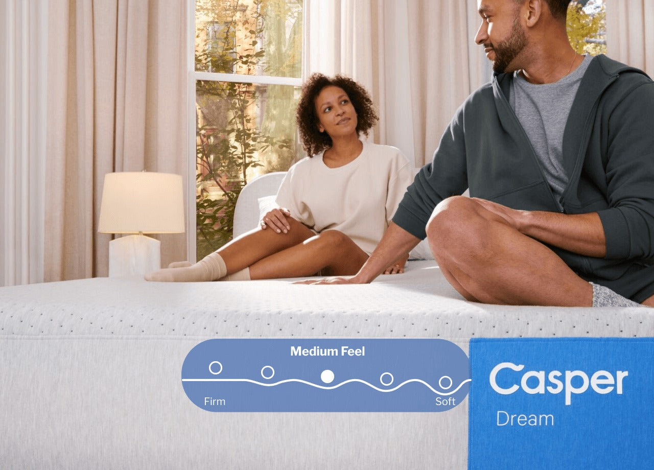 Front-facing image of a Casper Dream mattress. A couple is seen sitting on their bed behind the name tag. A digital illustration placed on the image shows that this mattress has a Medium feel between firm and soft.