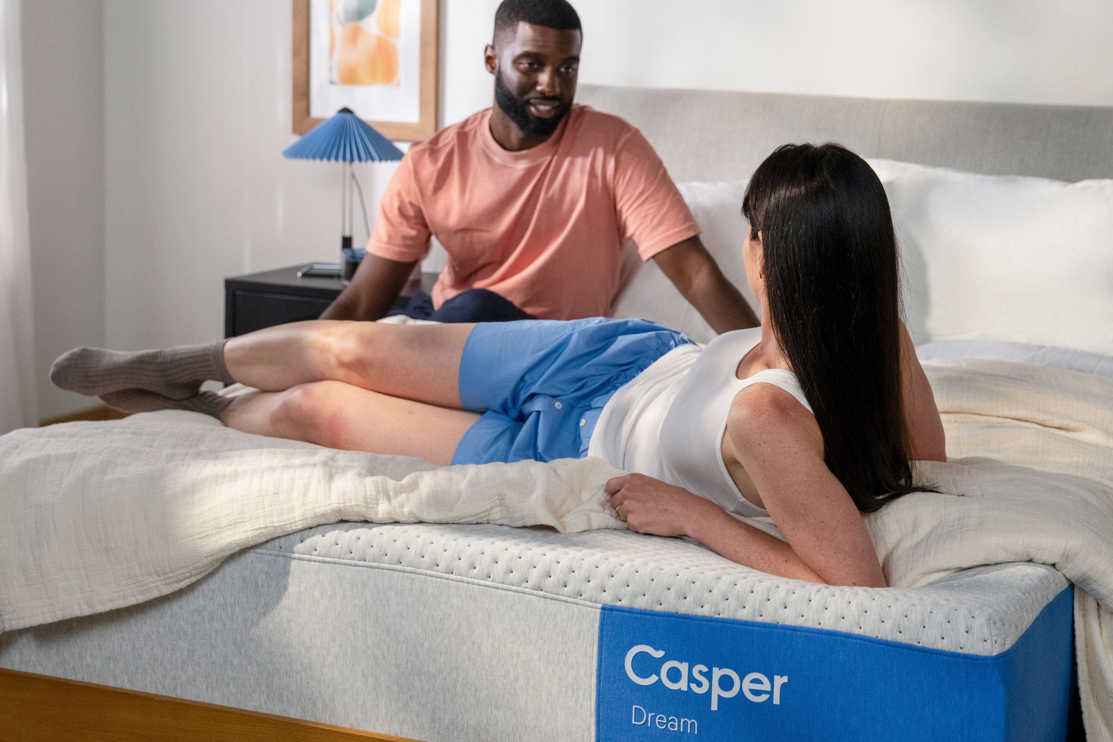 Image of a couple in bed on their Casper Dream mattress in a bedroom. One of them is lying back, sitting up on her elbows, and her partner is sitting on the edge of the bed and looking back at her.