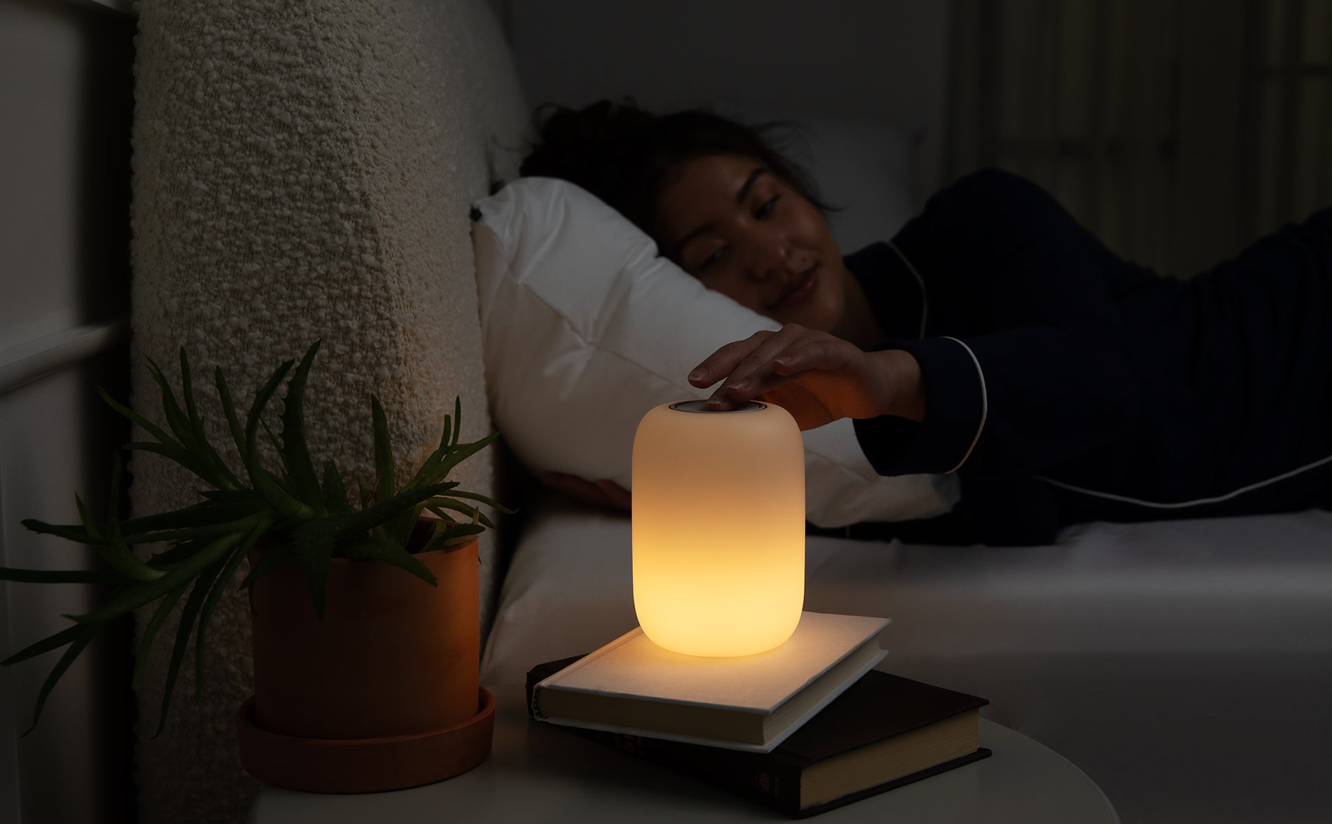 A Glow Light on a table table. A person lying in bed touching the power button.