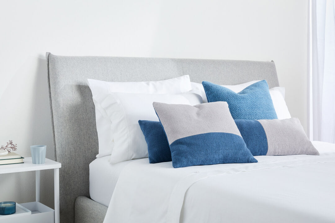 Light colors. A made bed with several pillows on it and a headboard against an off-white wall.