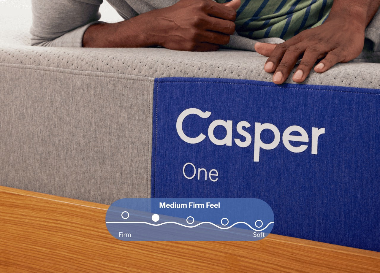 Close-up of the front side of a Casper One mattress. A person is seen resting on the mattress behind the name tag. A digital illustration placed on the image shows that this mattress has a Medium Firm feel, slightly more firm than it is soft.