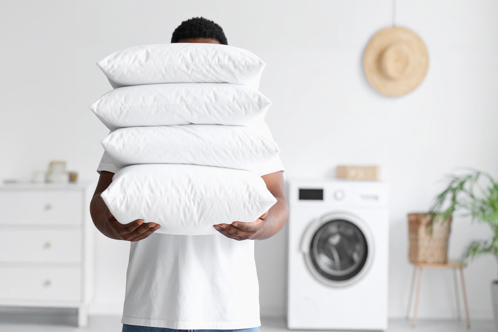 Person holding a stack of pillows in a laundry room with a washing machine in the background.