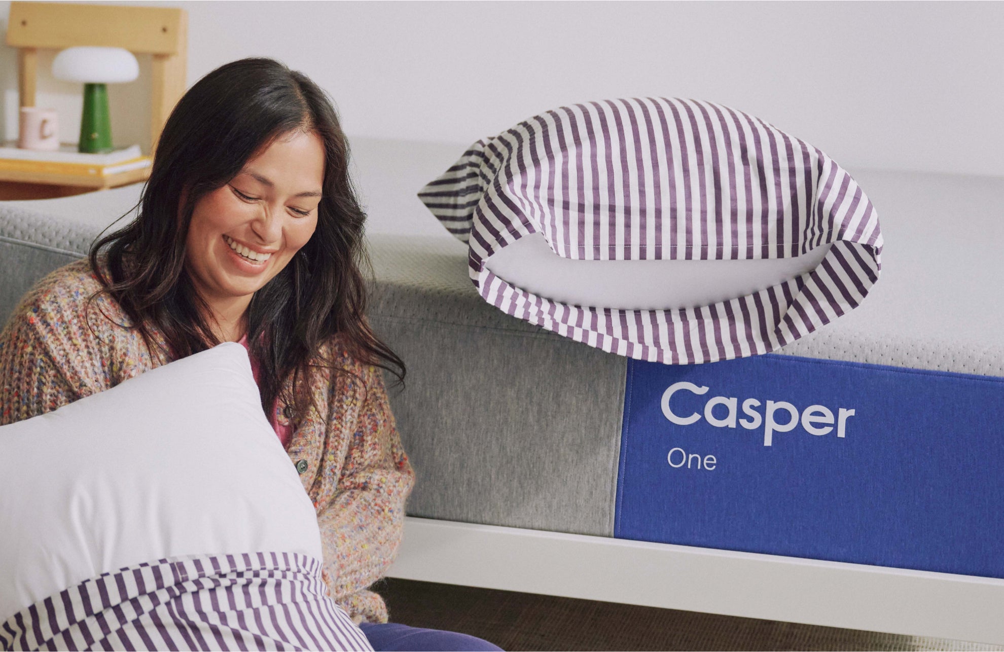 Woman sitting on a Casper mattress with a striped pillow, smiling.