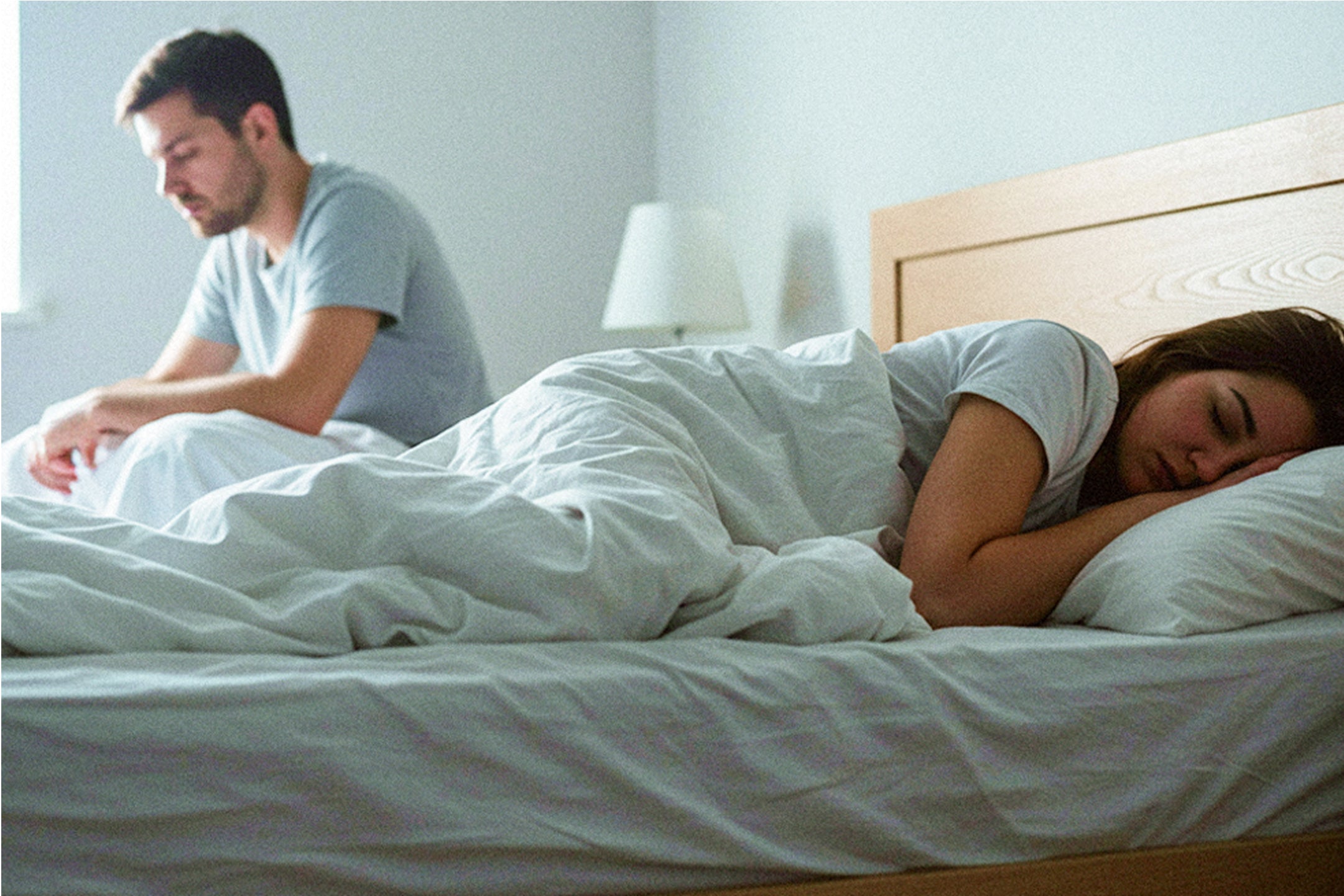 Side-view image of a couple in bed. A woman lies on her side sleeping. Next to her in the background, a man is sitting upright in bed.