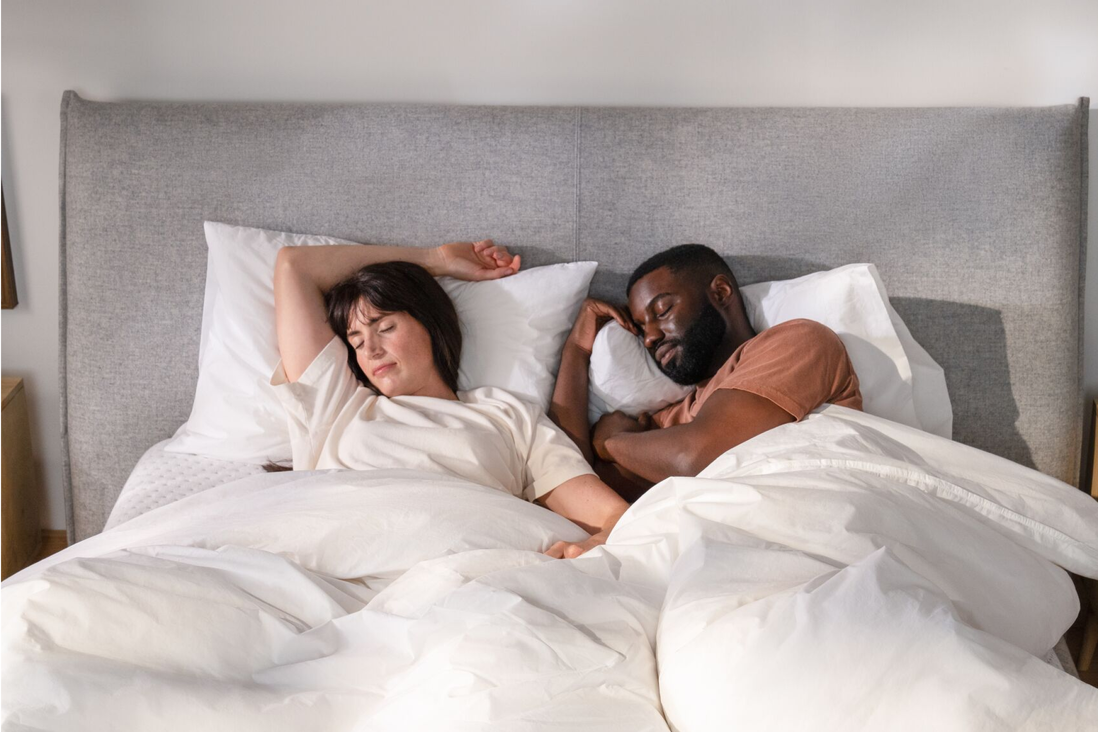 Front-facing image of a couple sleeping peacefully in bed. They are under their covers, lying on their pillows, which are leaning on a large headboard.