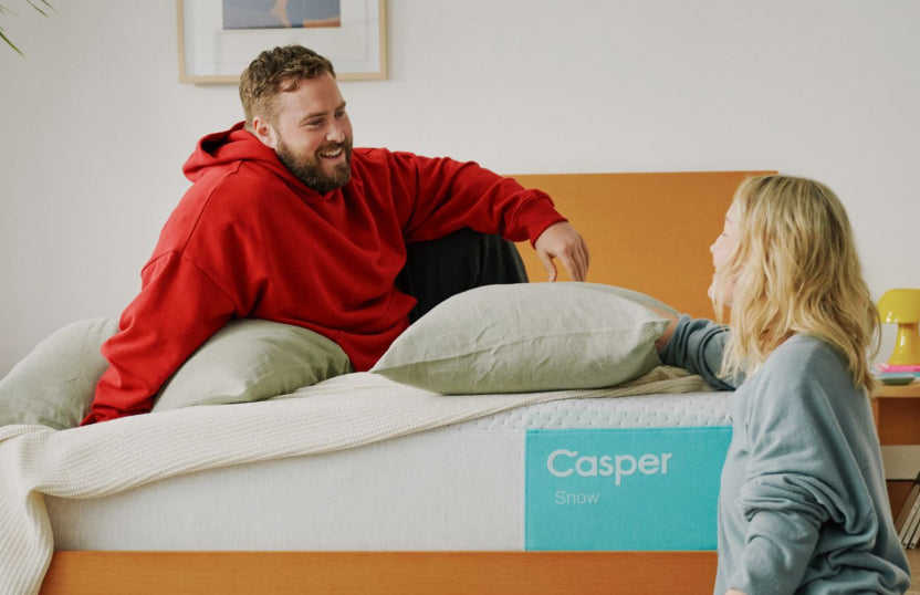 Two people sitting on and next to a Casper Snow mattress in a bedroom.