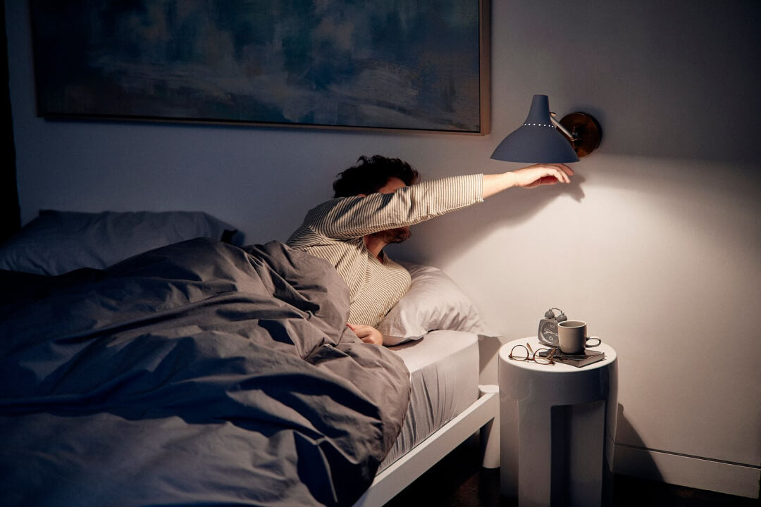 A person lying in bed at night reaches over to turn off the lamp.