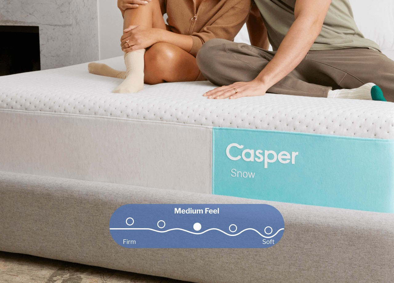 A couple sits on top of their Casper Snow mattress. A digital illustration placed on top of the image shows that this mattress has a Medium feel, between firm and soft.