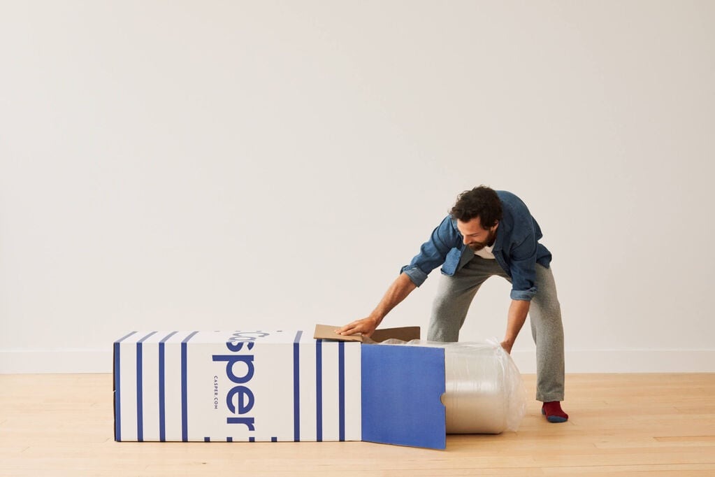A person unboxes a Casper mattress. It is in an air-tight bag. The box has Casper's blue and white logo on it.