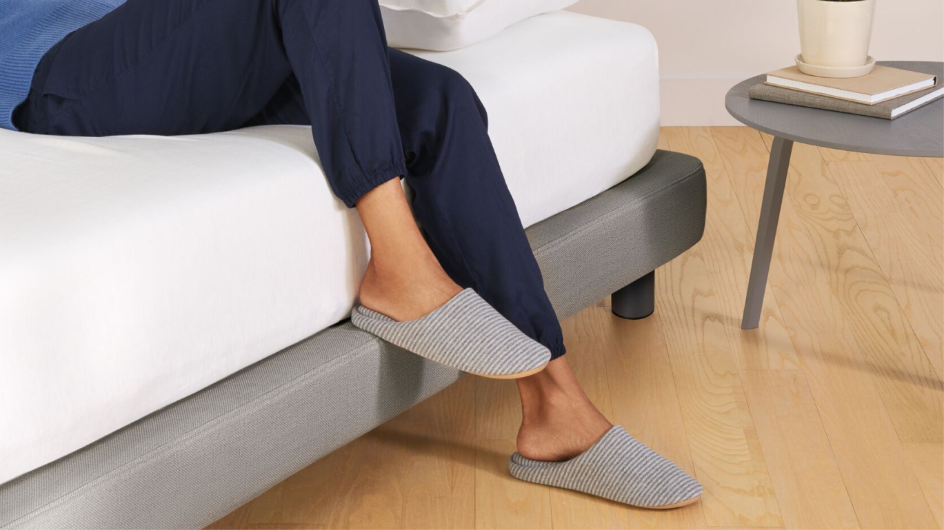 A person sitting on the edge of a bed with one foot on the floor and the other on the 2" ledge of the Upholstered Bed Frame, gray color.