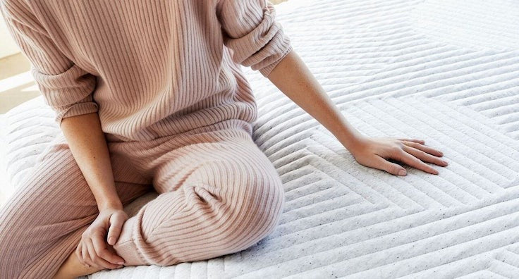 A person sits on a mattress and places their hand next to them.