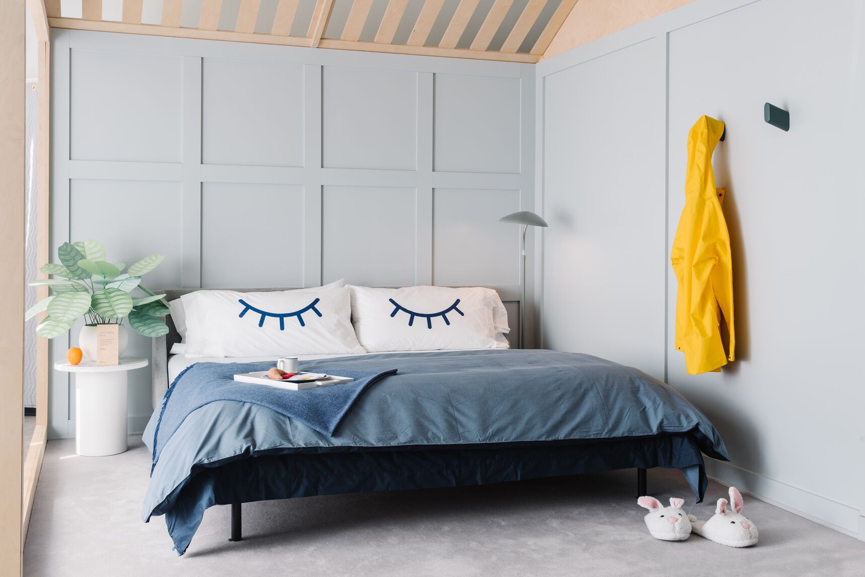 A bedroom with a fully made bed in it. The pillowcases have a graphic illustration of two closed eyelids. A plant is on a nightstand next to the bed.