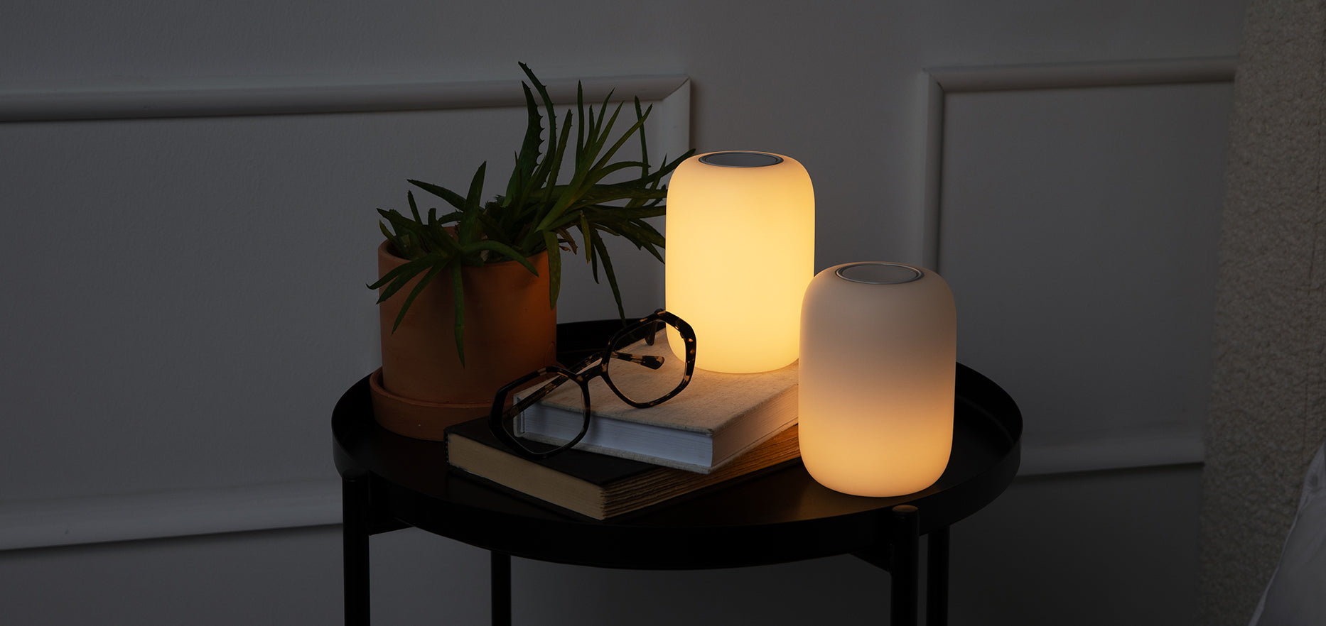 Lights-off image of two glow lights sitting on a night table with books and a plant