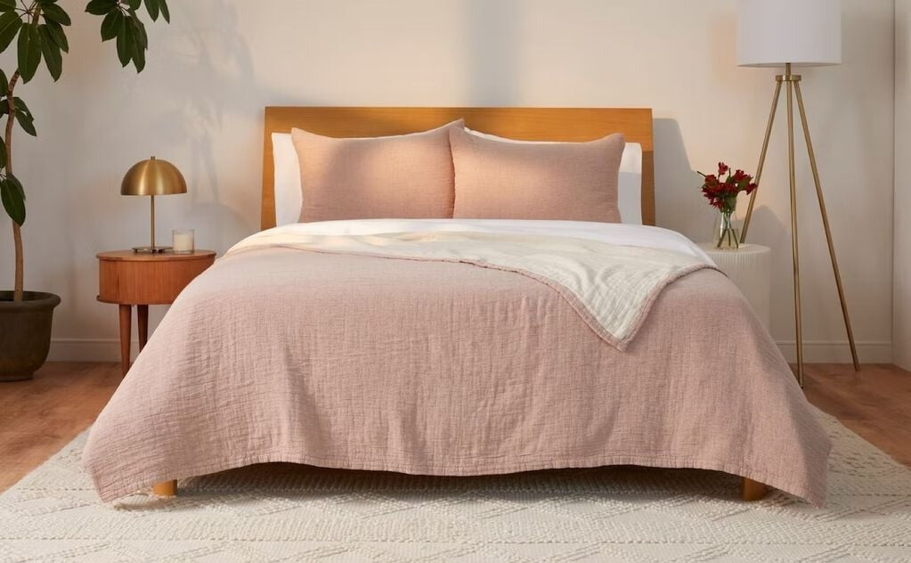 A bed with a spice colored matelasse coverlet, matching pillows, and a wooden headboard, set in a room with a side table, lamp, and plant.