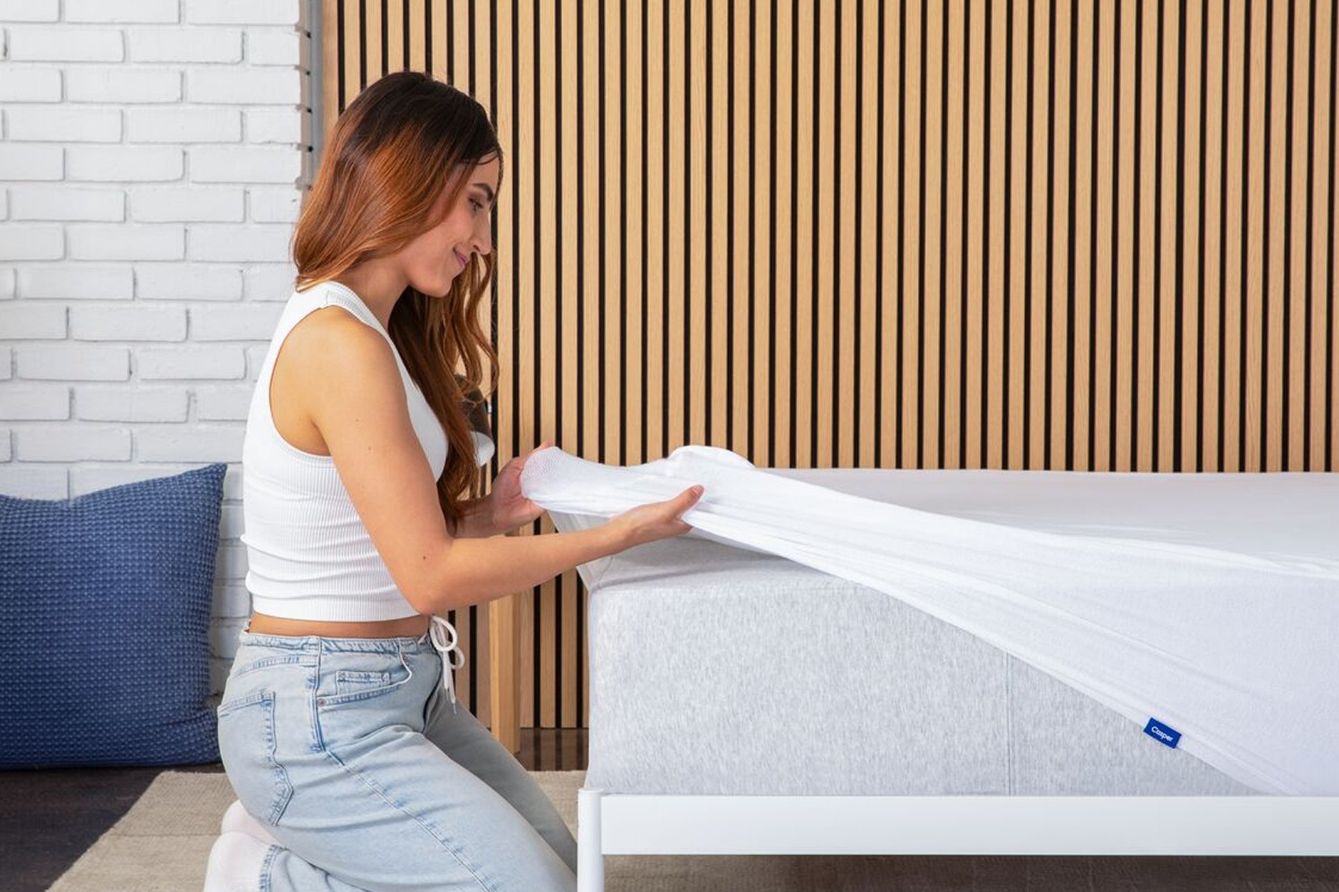Side view of a woman unrolling a mattress protector on a mattress in a bedroom setting.