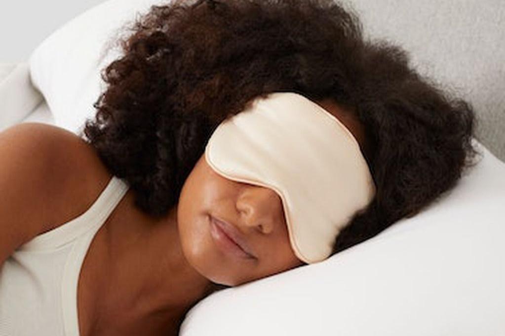 A woman wearing the Silk Sleep Mask, peach, in bed
