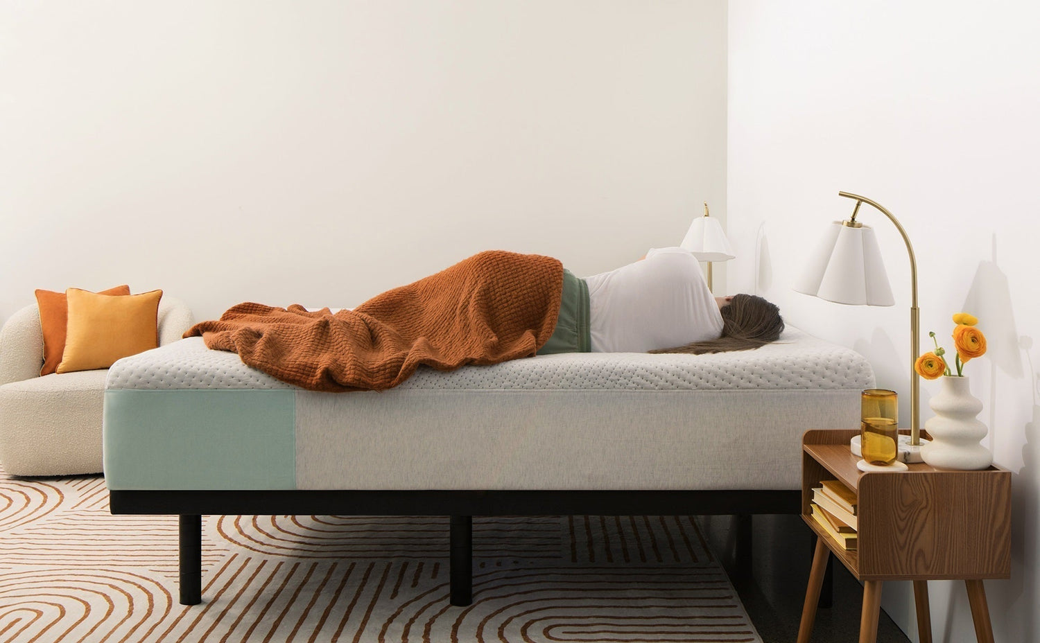 Young woman sleeping on their side on a memory foam mattress with a brown blanket in a cozy, modern bedroom featuring warm tones and minimalist decor.