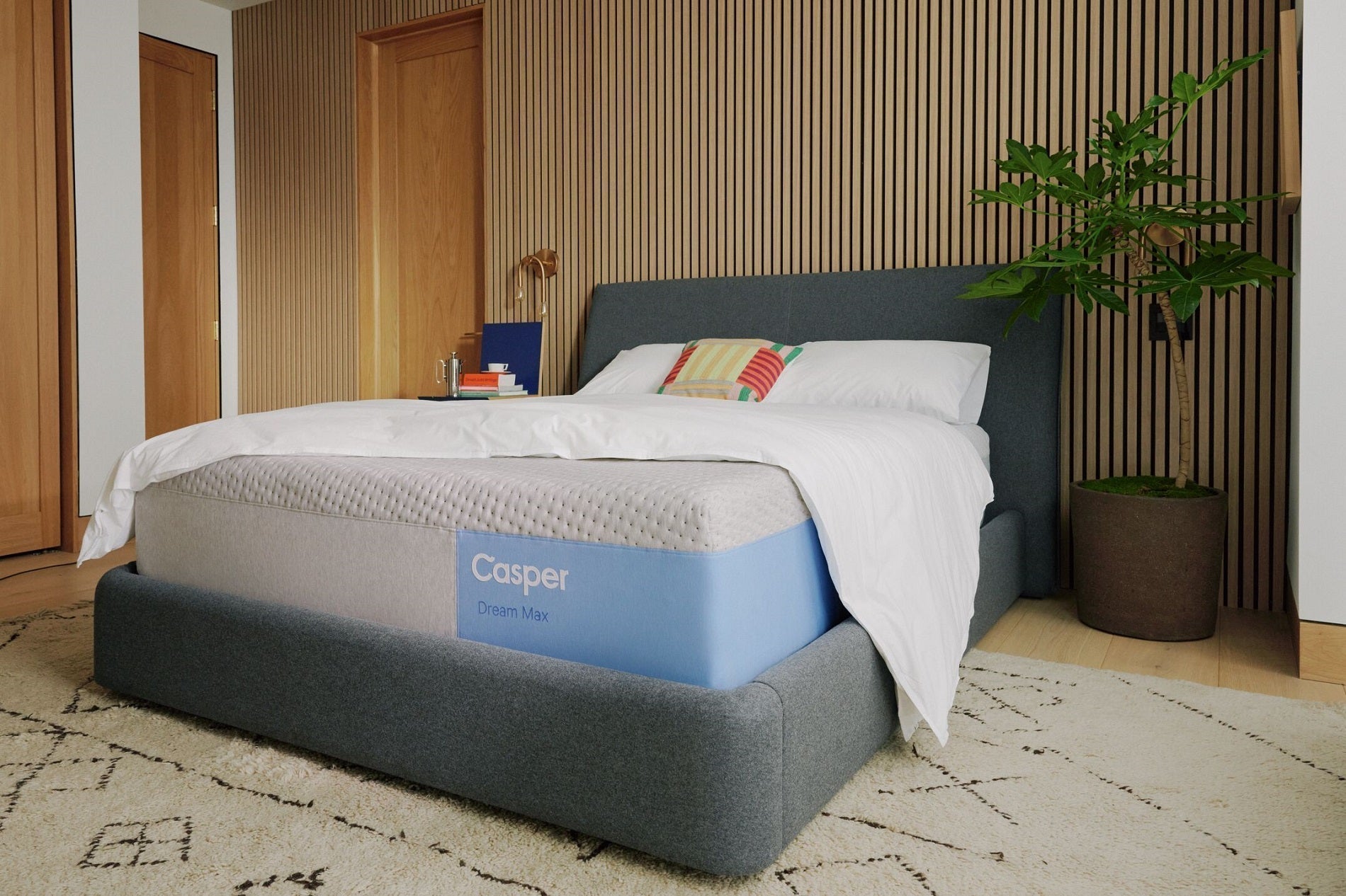  A close-up of a neatly made bed featuring a Casper Dream Max mattress on a gray upholstered frame in a modern bedroom with vertical wood paneling and a potted plant.