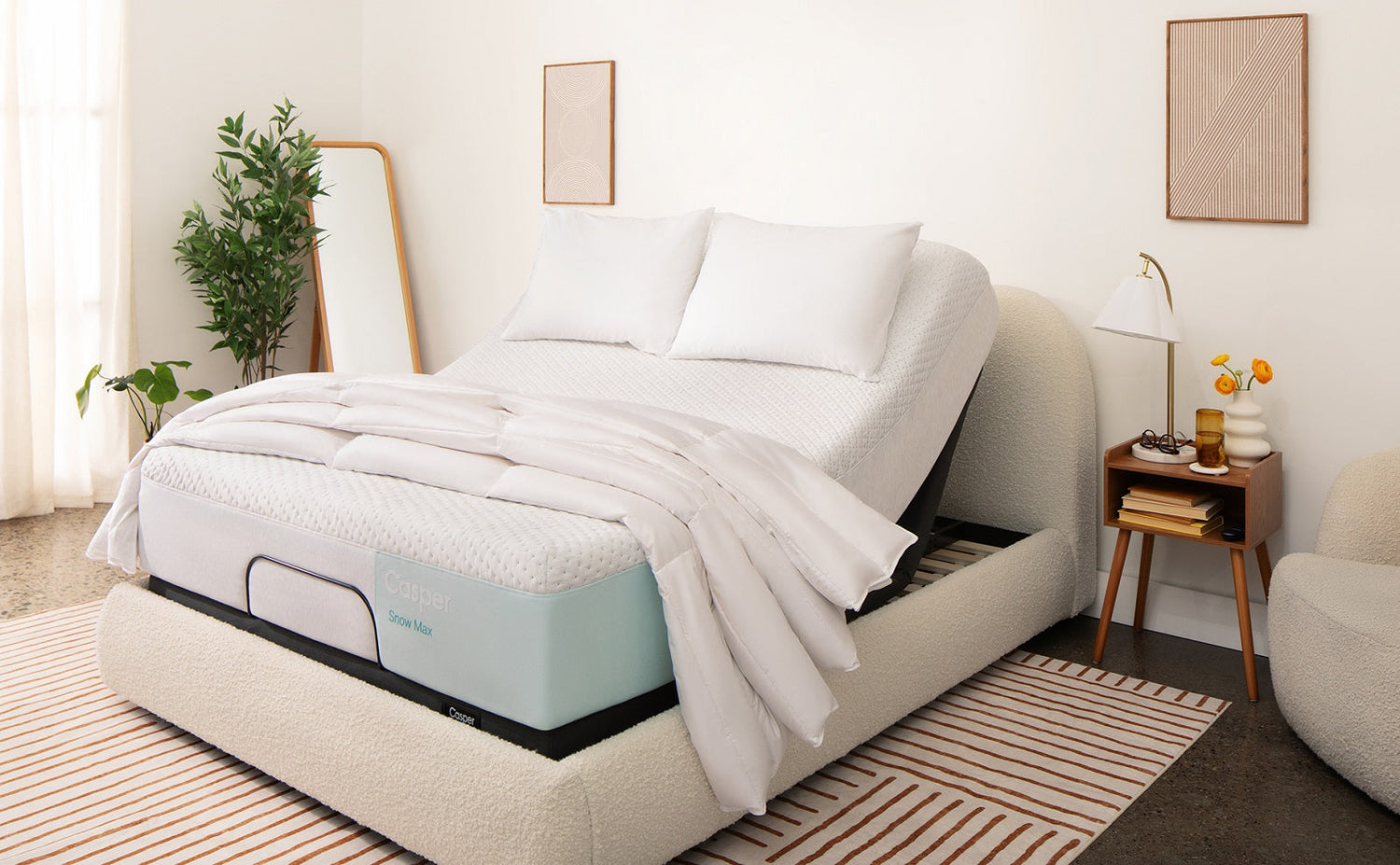 A white Casper Snow Max mattress is shown on an adjustable bed base in a bright, modern room. The bed features a textured cream headboard and frame, set against a striped rug and surrounded by minimalist decor.