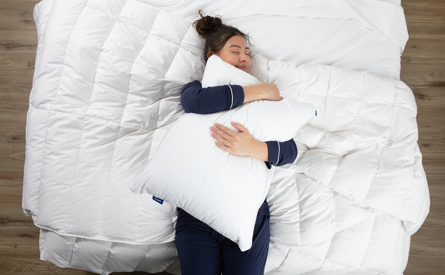 A woman in navy pajamas lies on a bed hugging a white Casper pillow, surrounded by soft white bedding.