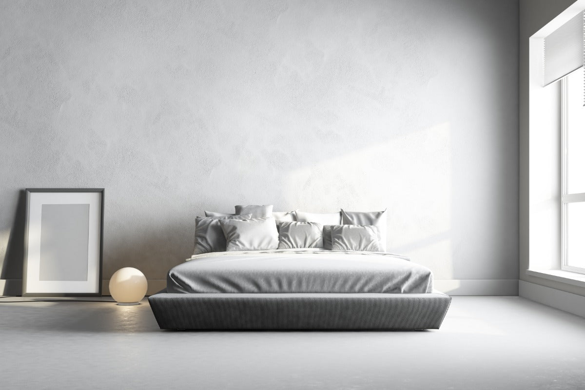 A low-profile platform bed with neatly arranged pillows centered in a spacious, minimalist bedroom with light grey walls and natural light from a large window.