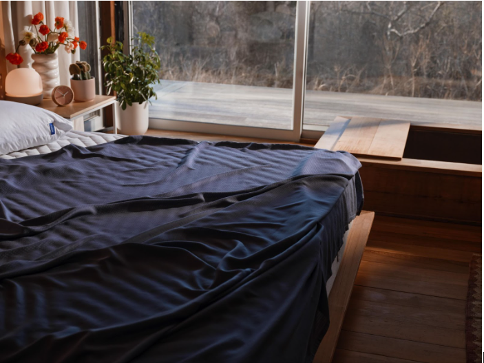 Large oversized bed with a dark blue sheet set in a sunlit bedroom next to floor-to-ceiling windows.