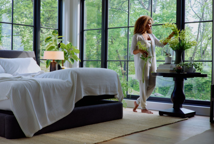 Woman arranging flowers by a large window in a bright bedroom next to a bed dressed in crisp white linens.